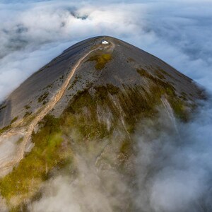 May include: An aerial view of a mountain peak shrouded in clouds. The mountain is green and rocky, with a winding path leading to the summit. The clouds are white and fluffy, creating a dramatic contrast to the dark green of the mountain.