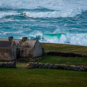 May include: A small, gray, two-story house with a stone wall in front of it. The house is on a grassy hill overlooking a stormy ocean with large waves crashing against the shore.