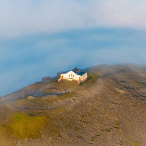 Könnte beinhalten: Luftaufnahme eines kleinen, weißen Gebäudes mit dunklem Dach auf einem Berggipfel. Das Gebäude hat eine rote Tür und Verkleidung. Der Berg ist von Nebel umhüllt, mit blauem Himmel darüber.