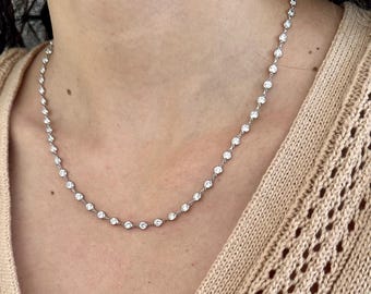 May include: A delicate silver necklace featuring a series of round, clear gemstones. The necklace is worn around the neck, showcasing the sparkling stones against the skin. The chain is thin and the gemstones are evenly spaced.