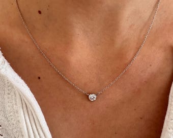 May include: A delicate silver necklace featuring a single, round, clear gemstone pendant. The pendant is centered on a fine chain, resting against a person's skin. The necklace is a simple, elegant piece of jewelry.