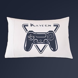 May include: White pillowcase with a navy blue graphic of a video game controller inside a triangle, with the name "KAYDEN" above it. The pillowcase is rectangular and appears to be made of soft fabric.