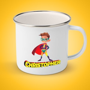 Op de afbeelding: Witte emaille mok met een cartoonillustratie van een superheldenjongen die een rode cape, een geel en blauw pak en een groene bril draagt. De tekst "CHRISTOPHER" is in gele letters onder de illustratie gedrukt.