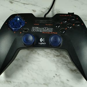 May include: A black Logitech WingMan RumblePad gaming controller featuring a blue directional pad, two blue analog sticks, and action buttons labeled A, B, C, and X. The controller includes a wired connection and text reading WingMan RumblePad.