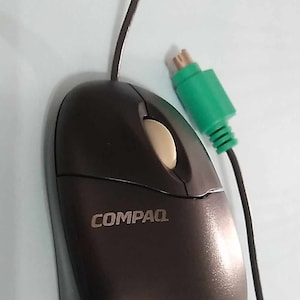 May include: A black Compaq computer mouse with a beige scroll wheel and a green connector. The mouse has a curved design and the Compaq logo is visible on the top. A black cable extends from the mouse.