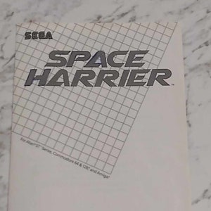 Peut inclure: Une jaquette de jeu vidéo Space Harrier en noir et blanc. La jaquette présente le titre du jeu en caractères gras blancs. Le texte "For Atari ST Series, Commodore 64 & 128 and Amiga" est imprimé en caractères gris plus petits en bas de la jaquette. La jaquette comprend également une description du jeu : "Blast incredibly evil, alien invaders into oblivion. Dodge deadly obstacles. And restore peace to the beautiful Land of Dragons."
