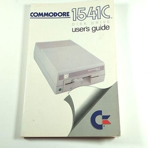 May include: A vintage Commodore 1541C disk drive user's guide. The cover features a graphic of the disk drive and the Commodore logo. The guide is white with blue and red text. The disk drive is light grey.