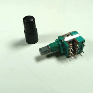 May include: A rotary potentiometer with a green plastic housing and a metal shaft, accompanied by a separate cylindrical black plastic knob. The electronic component features multiple metal pins extending from the rear for circuit board connection.