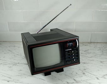 May include: A vintage black and red portable television with a silver antenna. The TV has a small screen and a control panel on the side. The TV is sitting on a white countertop.