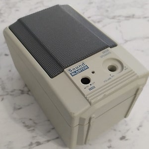 May include: A beige Sound Blaster speaker with a grey speaker grille, a volume knob, and an on/off switch. The speaker has a black and blue Sound Blaster logo.