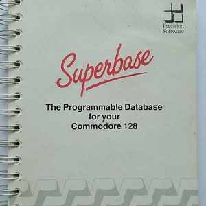 Puede incluir: Un libro blanco con espiral y encuadernación negra. La portada tiene un logotipo rojo que dice "Superbase" y el texto "The Programmable Database for your Commodore 128". El logotipo de Precision Software está en la esquina superior derecha.