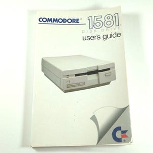 May include: A vintage Commodore 1581 disk drive and user's guide. The guide is white with blue text and a graphic of the disk drive. The disk drive is beige and rectangular. The Commodore logo is visible.