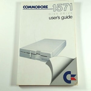 May include: A vintage Commodore 1571 Disk Drive user's guide. The cover features a graphic of the disk drive and the Commodore logo. The guide is white with black and blue text.
