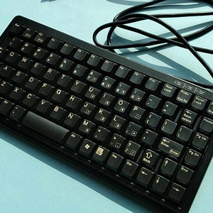 Könnte beinhalten: Eine schwarze, kabelgebundene Tastatur mit Standardlayout und goldenen Beschriftungen. Die Tastatur befindet sich auf einer hellblauen Oberfläche.