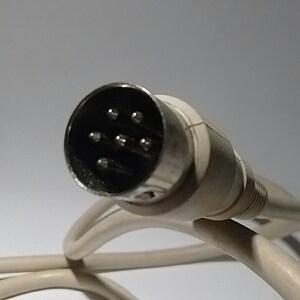 May include: A white circular DIN connector with five metal pins arranged in a semi-circular pattern, attached to a flexible white cable. The connector features a metallic outer ring and a moulded white plastic housing that tapers towards the cord.