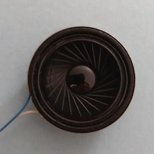 May include: A black round speaker with a black wire attached. The speaker has a black cone with a circular pattern of lines radiating from the center.