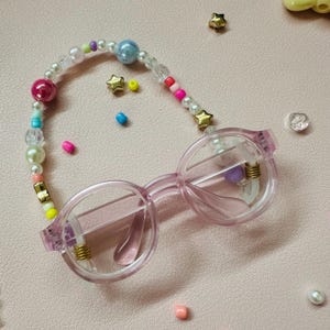 May include: A pair of light pink round eyeglasses with a beaded eyeglass chain. The chain features a variety of colorful beads, including pearls, gold star-shaped beads, and other colorful beads. The glasses are on a pink surface.