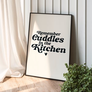 Op de afbeelding: Een zwart ingelijst print met een witte achtergrond en zwarte tekst die "Remember cuddles in the kitchen" luidt met een klein hartje onder de tekst.