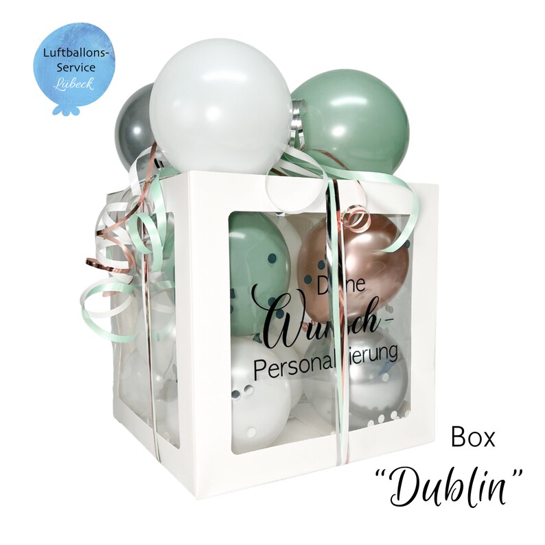 May include: A white box with a clear window filled with balloons. The balloons are green, white, silver, and rose gold. The box has a ribbon tied around it and the text "Die Wunsch-Personalisierung" is printed on the box. The box is labeled "Box "Dublin"".