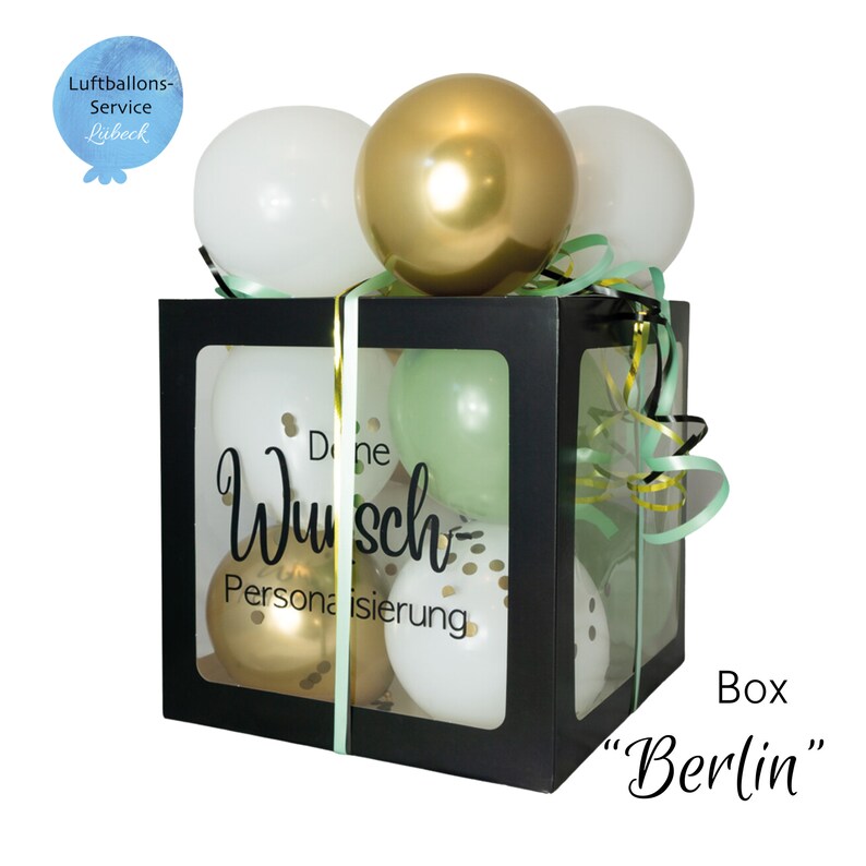 May include: A black box with a clear window filled with balloons. The balloons are white, gold, and green. The box has a ribbon tied around it and the text "Deine Wunsch Personalisierung" is printed on the front. The box is labeled "Box "Berlin"".