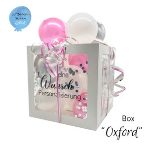 Op de afbeelding: Een witte doos met een transparant venster gevuld met roze, witte en zilveren ballonnen. De doos heeft een lint eromheen en de tekst "eine Wunsch-Personalisierung" staat op de zijkant gedrukt. De doos is gelabeld "Box "Oxford"".