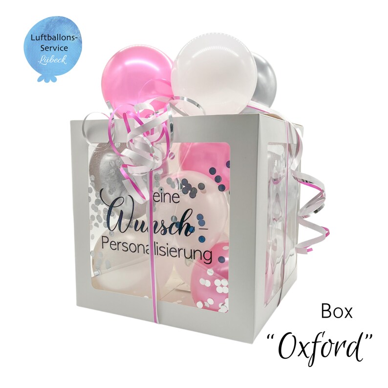 Op de afbeelding: Een witte doos met een transparant venster gevuld met roze, witte en zilveren ballonnen. De doos heeft een lint eromheen en de tekst "eine Wunsch-Personalisierung" staat op de zijkant gedrukt. De doos is gelabeld "Box "Oxford"".