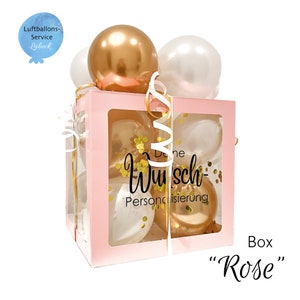 May include: A pink box with a clear window filled with gold and white balloons. The box has a gold ribbon and confetti inside. The text "Deine Wunsch-Personalisierung" is printed on the box.
