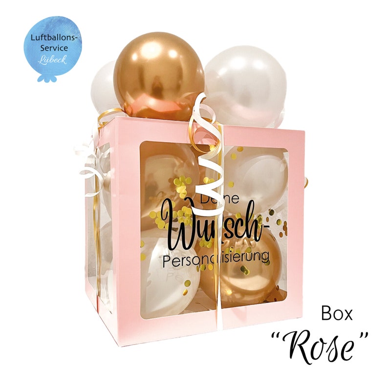 May include: A pink box with a clear window filled with gold and white balloons. The box has a gold ribbon and confetti inside. The text "Deine Wunsch-Personalisierung" is printed on the box.