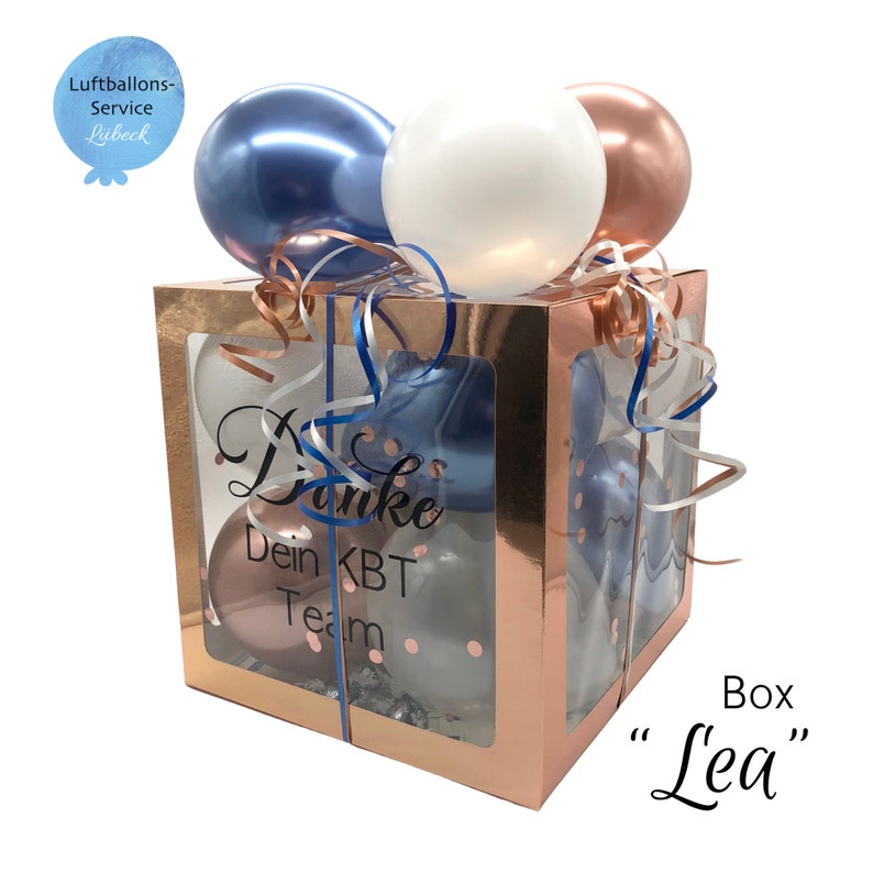 May include: A rose gold gift box with a clear window, filled with blue, white, and rose gold balloons. The box has a ribbon tied around it and the text "Danke Dein KBT Team" is printed on the side. The box is labeled "Box "Lea"".