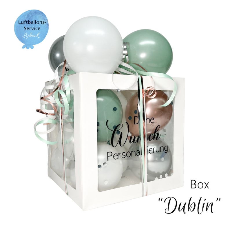 Op de afbeelding: Een witte doos met een doorzichtig venster gevuld met ballonnen. De ballonnen zijn groen, wit, zilver en ros&eacute; goud. De doos heeft een lint eromheen gebonden en de tekst "Die Wunsch-Personalisierung" staat op de doos gedrukt. De doos is gelabeld met "Box "Dublin"".