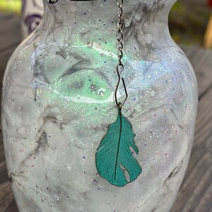 May include: A clear glass vase with a white and gray marbled design and iridescent glitter. The vase has a silver mesh tea infuser with a teal feather charm hanging from it.