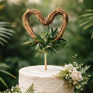 May include: A two-tier cake with off-white textured frosting, decorated with fresh green ferns, white baby's breath, and pale pink roses. A wooden stick supports a heart-shaped topper made of intertwined brown twigs and green leaves.