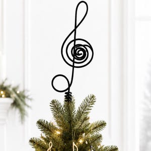 May include: A black metal treble clef tree topper sits atop a green Christmas tree decorated with white and black ornaments featuring musical notes, gold miniature instruments, and warm white string lights throughout the evergreen branches.