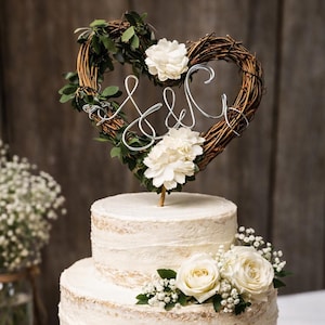 May include: A two-tier white frosted cake decorated with white roses and baby's breath. A heart-shaped twig wreath topper features green leaves, white flowers, and silver wire script letters S & C, all resting on a round wooden slice base.
