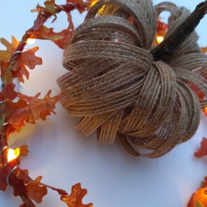 Op de afbeelding: Een handgemaakte pompoen van jute met een takje als steel, omringd door een slinger van oranje herfstbladeren en kleine, warmwitte lampjes. Een feestelijke herfstdecoratie.