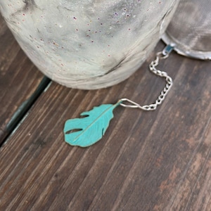 Könnte beinhalten: Ein blaugrüner Metall-Tee-Infuser in Form einer Feder mit einer silbernen Kette, die an einem Sieb befestigt ist.