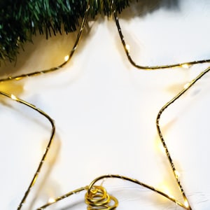 Könnte beinhalten: Ein goldener Drahtstern mit weißen Lichtern, die um ihn herumgewickelt sind. Der Stern hängt an einer goldenen Spirale.
