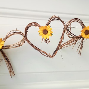 Op de afbeelding: Een decoratieve krans gemaakt van bruine takken, gevormd in twee harten met gele zonnebloem accenten.