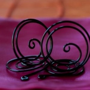 Può includere: Due anelli per tovaglioli in metallo nero con un design a spirale decorativo. Gli anelli sono su una superficie di tessuto rosa.