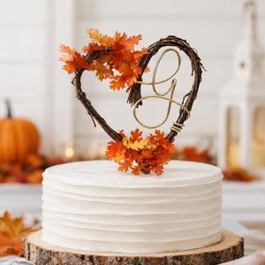 May include: A white frosted cake sits on a wood slice base, topped with a heart-shaped grapevine wreath adorned with orange and red autumn leaves and a gold wire script letter G. The cake is textured with horizontal ridges along the sides.