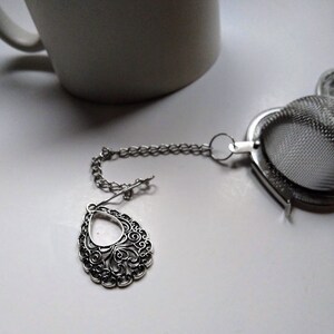 Może przedstawiać: Srebrny zaparzacz do herbaty o delikatnym, ozdobnym wzorze. Zaparzacz jest przymocowany do łańcuszka i ma siateczkową kulkę do luźnej herbaty.