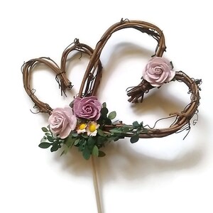 Op de afbeelding: Een rustieke taarttopper met twee in elkaar grijpende hartvormen gemaakt van bruine twijgen. De harten zijn versierd met lichtroze en mauve stoffen rozen, kleine gele bloemen en groene bladeren. Een houten deuvel loopt van de onderkant.