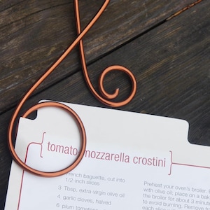May include: Copper-colored metal recipe holder in the shape of a treble clef, holding a recipe card. The card features the title "tomato mozzarella crostini" and a list of ingredients and instructions. The holder is on a dark wooden surface.