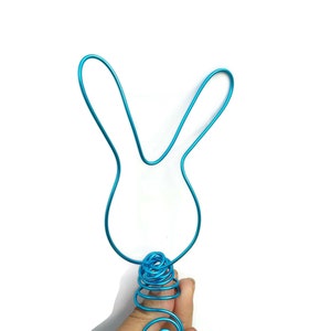 May include: A turquoise wire bunny head with long ears and a decorative spiral base. The wire is bent into the shape of a bunny, perfect for Easter or spring decor. The bunny is held by a hand against a white background.