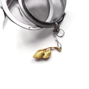 May include: A silver mesh tea infuser with a gold leaf-shaped chain attached. The infuser is designed to hold loose tea leaves and is perfect for brewing tea in a cup or teapot.