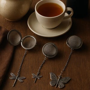Peut inclure: Nature morte avec une tasse de thé, une théière et un récipient noir. Trois infuseurs à thé avec des breloques libellule et papillon sont également présents. Les infuseurs à thé sont argentés et ont un design en maille.