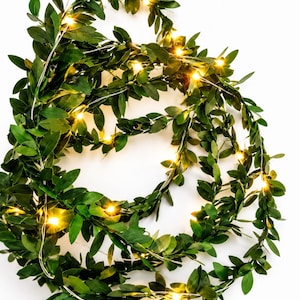 May include: A decorative garland featuring artificial green leaves intertwined with a thin silver wire string of small, warm white LED lights. The flexible vine-like strand is coiled in a circular shape, showcasing numerous pointed foliage.