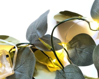 May include: A string of fairy lights with small, warm white bulbs wrapped around artificial eucalyptus leaves. The leaves are a mix of light and dark green.