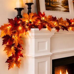 Puede incluir: Una guirnalda de otoño artificial con hojas de arce realistas en tonos naranja, rojo y amarillo. La guirnalda está entrelazada con pequeñas luces LED de color blanco cálido que emiten un brillo suave sobre el borde de la repisa blanca.
