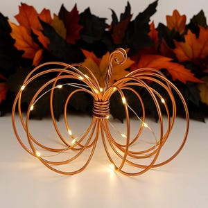 May include: A decorative pumpkin sculpture crafted from coiled copper-colored wire, featuring a central stem and rounded segments. The structure is illuminated by thin, integrated string lights that emit a warm glow throughout the wire frame.